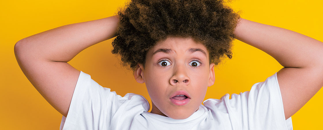 A boy holds both hands on top of his head with wide eyes and an open mouth